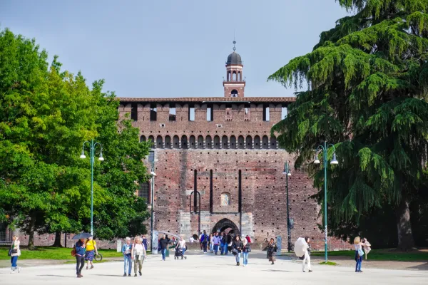 Gente paseando frente al Castillo Sforzesco en Milán, Italia, con árboles grandes enmarcando la vista.