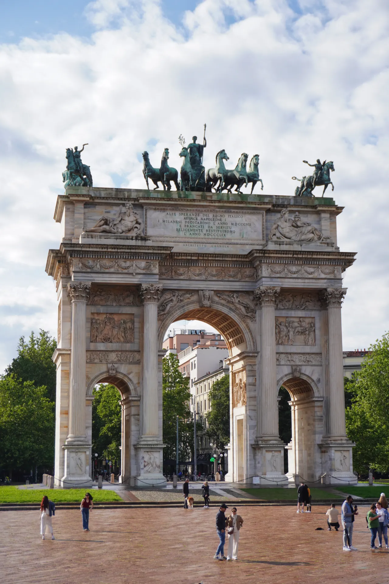 The imposing Arco della Pace in Milan, Italy, under a cloudy sky, with people walking in the foreground and trees surrounding the monument.