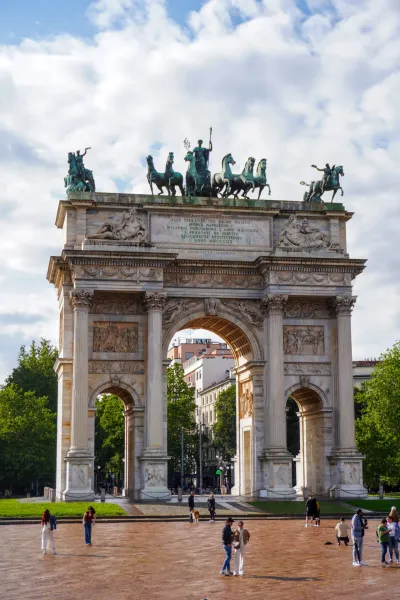 El imponente Arco della Pace en Milán, Italia, bajo un cielo nublado, con gente en primer plano y árboles rodeando el monumento.
