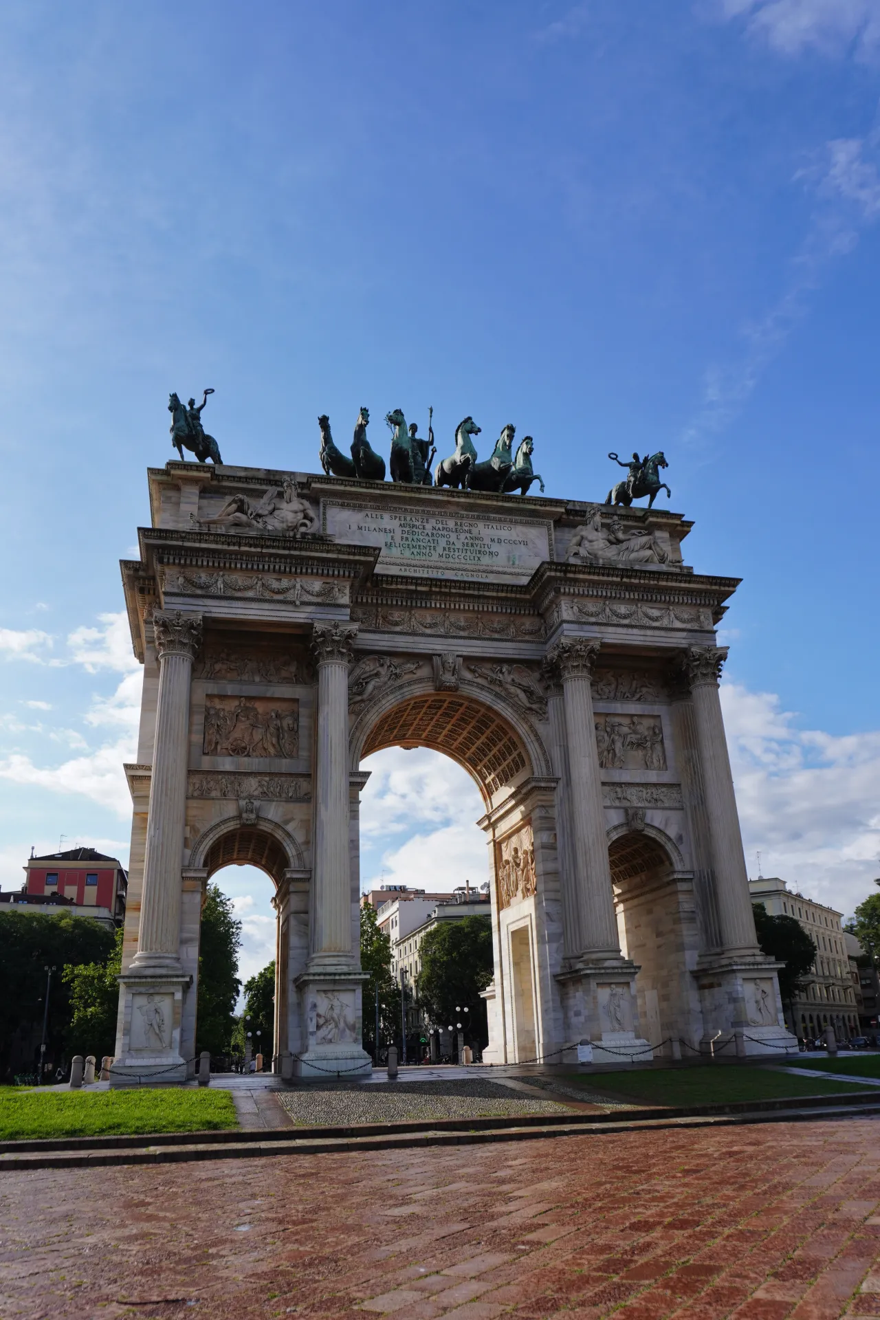 Arko mira, grandiozna neoklasična arkada u Milanu, Italija, sa skulpturom Kvadruge na vrhu, pod vedrim plavim nebom sa povremenim oblacima.