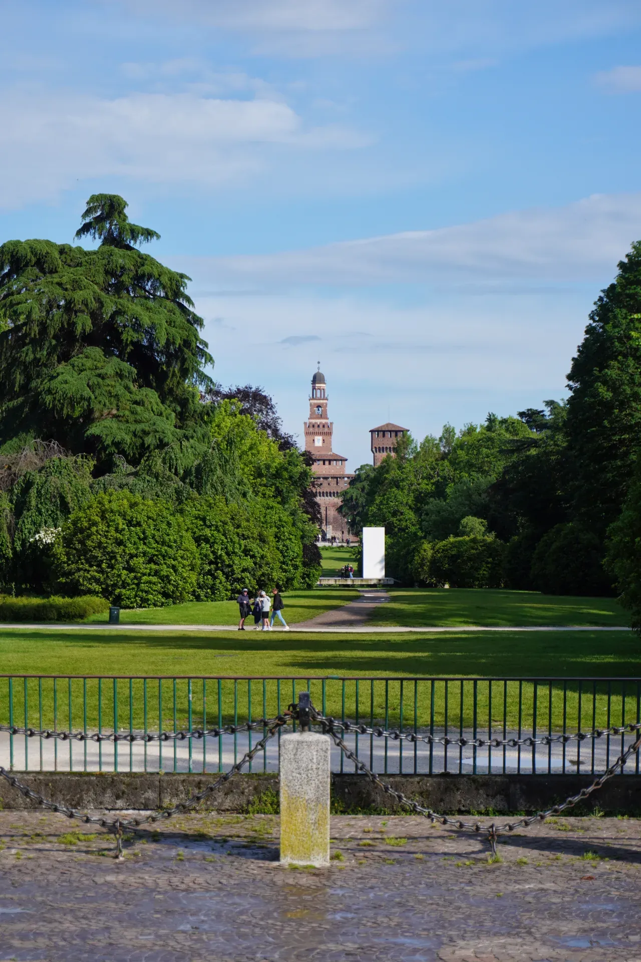 La tour du Château Sforza s'élève au-dessus des arbres dans le Parco Sempione, Milan, avec des personnes marchant sur un sentier au premier plan.