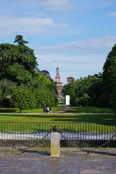 La torre del Castillo Sforza se eleva sobre los árboles en el Parco Sempione, Milán, con personas caminando por un sendero en primer plano.