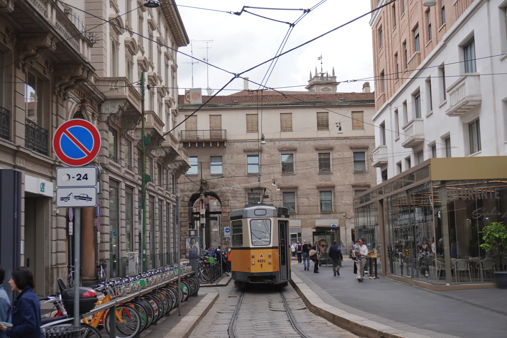 A yellow and beige tram travels down a street in Milan, Italy, with ornate historical buildings on one side and a modern glass-fronted restaurant on the other. Bicycles are parked along the sidewalk.