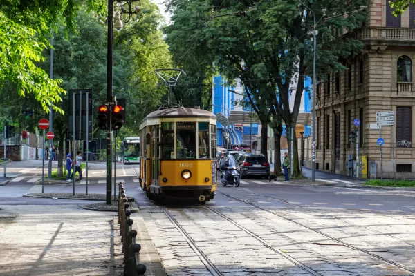 Un tranvía amarillo vintage, número 1602, circula sobre las vías por una calle de Milán, con árboles, edificios, un autobús, un coche y una motocicleta al fondo.