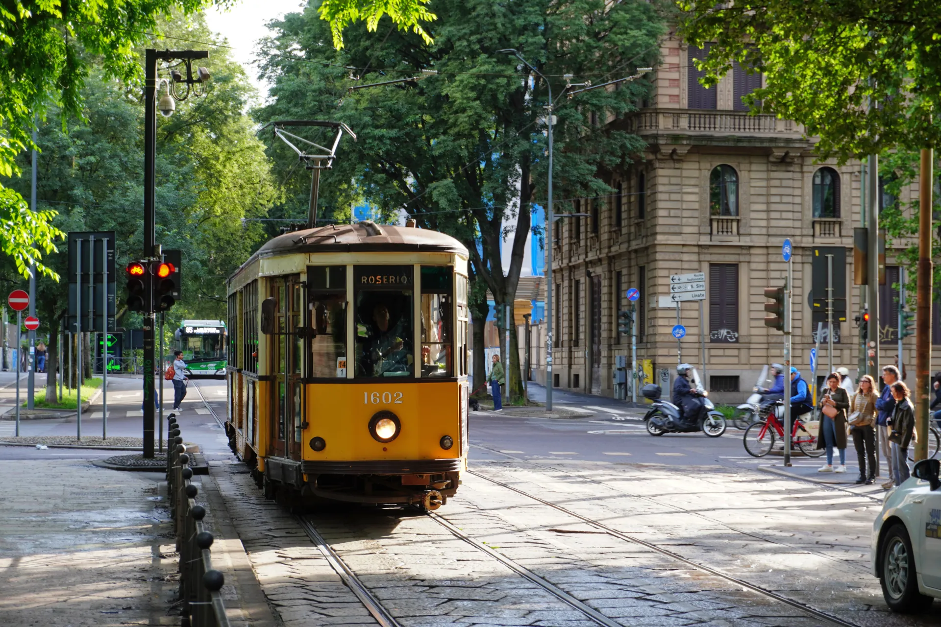 A vintage yellow tram, number 1602, moves along tram tracks on a street in Milan. The tram has 'ROSERIO' displayed on its front. In the background are buildings, trees, traffic lights, and people.