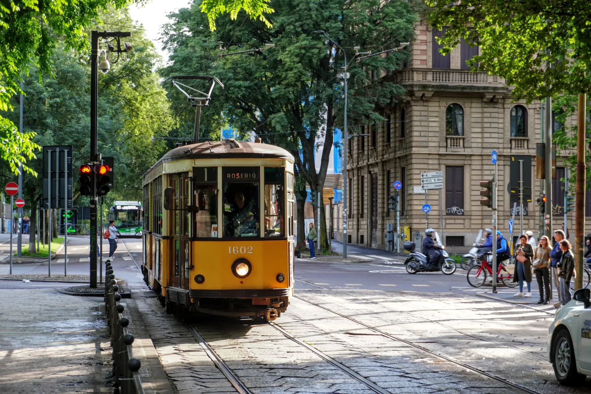 A yellow vintage tram, number 1602, moves along tracks on a street in Milan. The front of the tram displays 'ROSERIO'. In the background, buildings, trees, traffic lights, and people are visible.