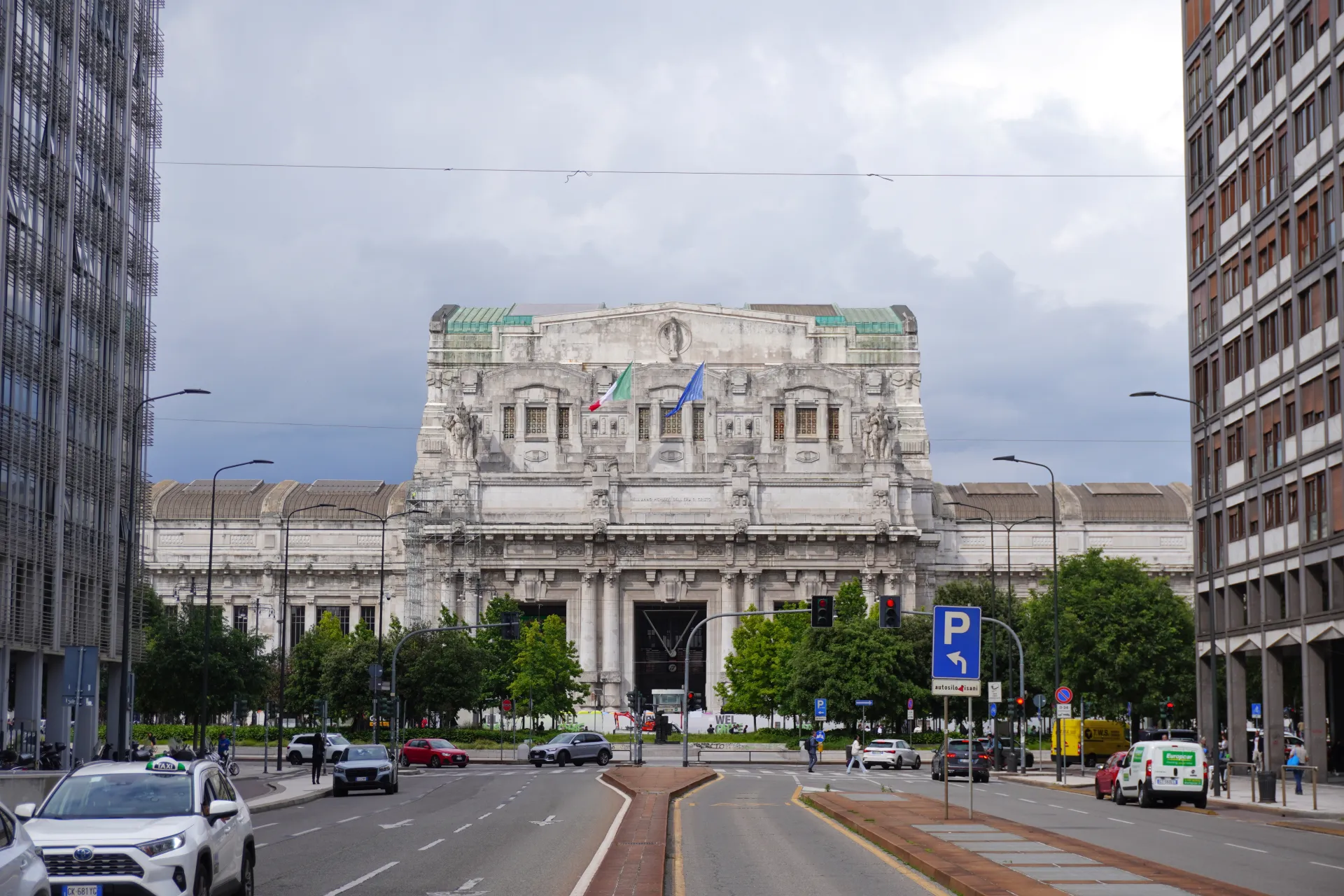 The impressive facade of Milan Central Station with Italian and European flags flying, surrounded by trees and with traffic on the street in the foreground.