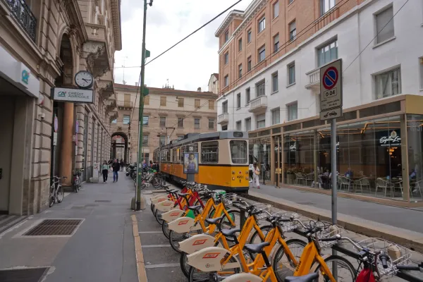 Eine Straße in Mailand, Italien, mit einer gelben Straßenbahn auf Gleisen, einer Reihe von orangen Leihfahrrädern, Fußgängern auf dem Bürgersteig und Gebäuden mit Geschäften und Cafés.