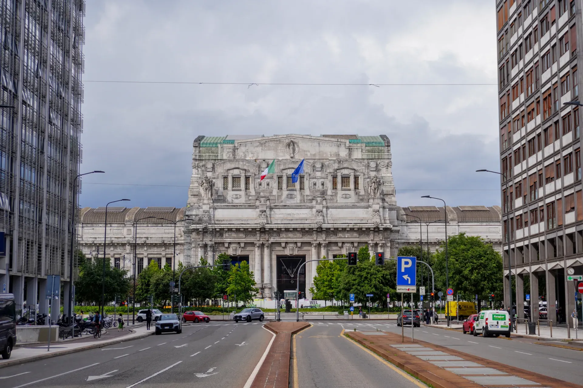 Die beeindruckende Fassade des Mailänder Hauptbahnhofs mit italienischen und europäischen Flaggen, umgeben von Bäumen und mit Verkehr auf der Straße im Vordergrund.