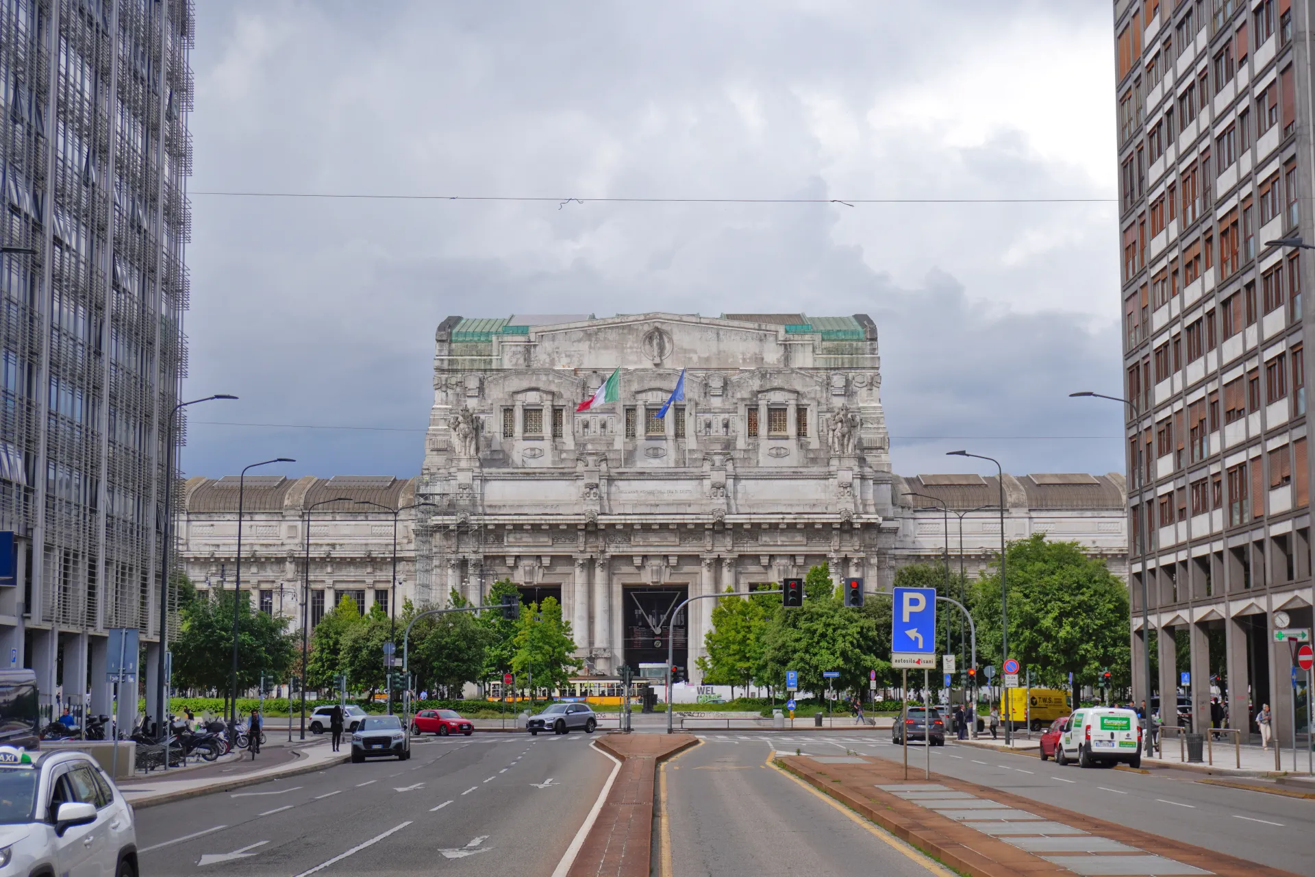 View of Milan Central Station with its elaborate facade, flags, modern buildings, street, cars, and trees under a cloudy sky.