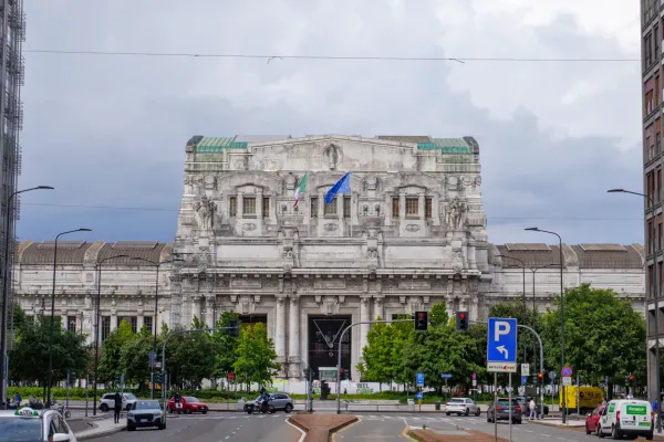 La imponente fachada de la Estación Central de Milán con banderas italianas y de la UE en un día nublado, con coches y árboles en la calle en primer plano.