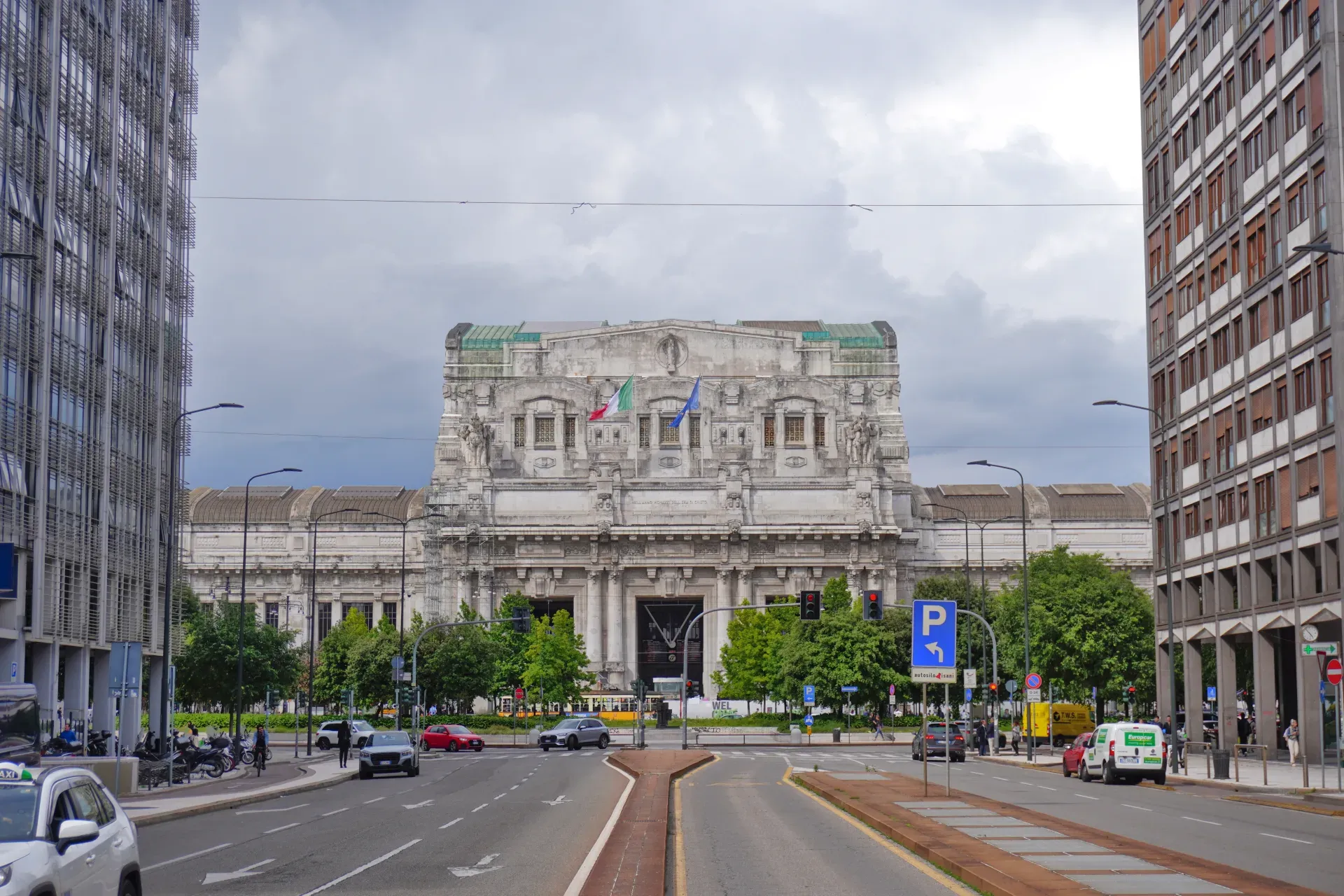 Vue de la gare centrale de Milan avec sa façade complexe, ses drapeaux, ses bâtiments modernes, sa rue, ses voitures et ses arbres sous un ciel nuageux.
