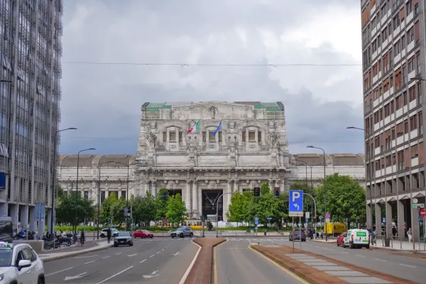 Vista de la Estación Central de Milán con su intrincada fachada, banderas, edificios modernos, calle, coches y árboles bajo un cielo nublado.