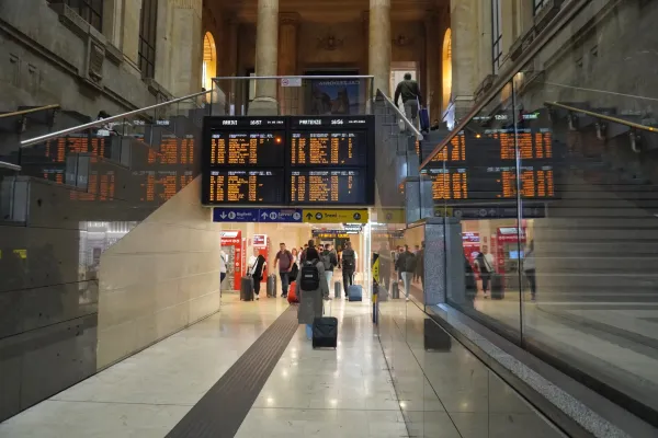 Viajeros con maletas en el gran vestíbulo de la Estación Central de Milán, con paneles electrónicos de salidas y llegadas que muestran información de trenes.