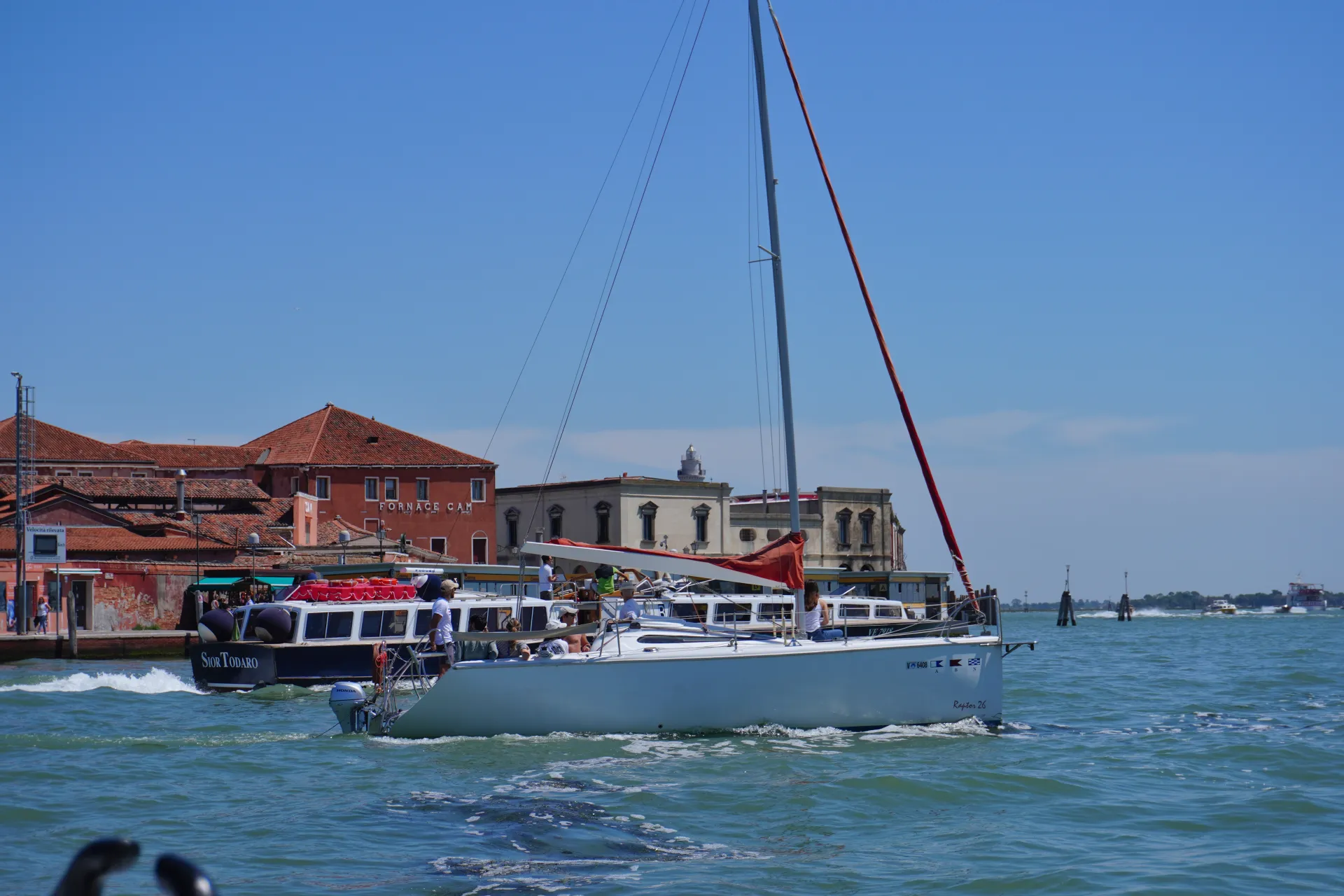 A white sailboat with a red sail navigating the turquoise waters of Venice, with a blue ferryboat and characteristic Venetian buildings in the background under a bright blue sky.