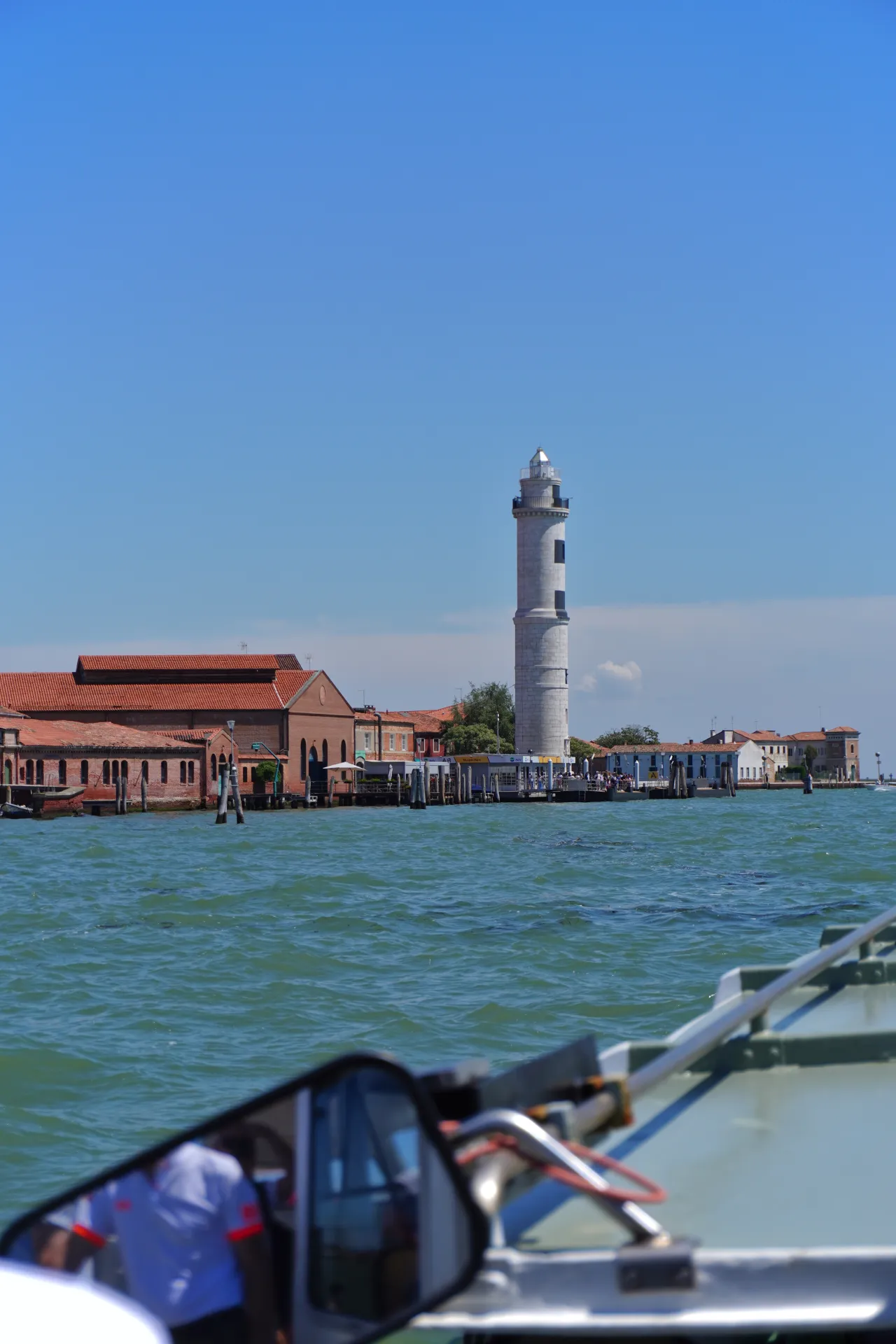 A tall, white lighthouse with dark windows stands prominently against a bright blue sky, with red-tiled buildings and a waterfront promenade to its left, all seen across a blue body of water. A boat's side mirror reflects a person wearing a white shirt in the foreground.