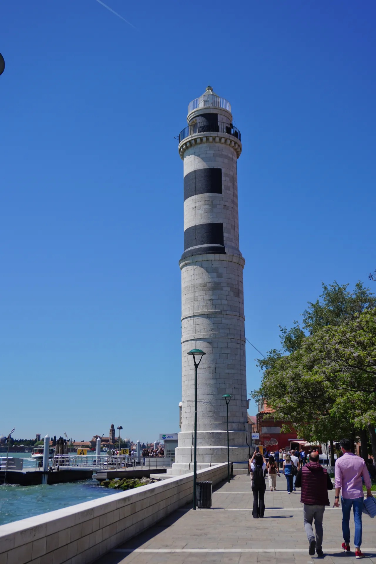 A tall, striped lighthouse on the Venice waterfront with people walking along the promenade and boats in the lagoon.