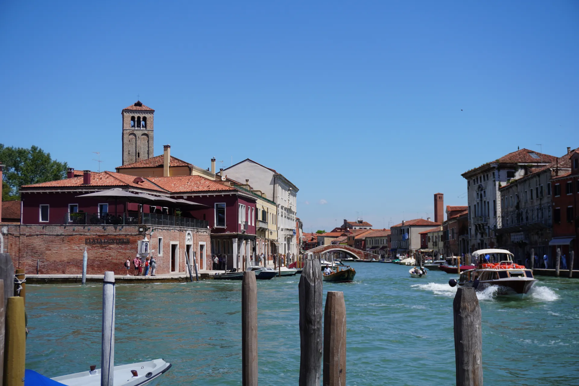 A sunny day in Venice, Italy, with boats moving along a canal, lined by colorful buildings and a prominent brick bell tower in the background.
