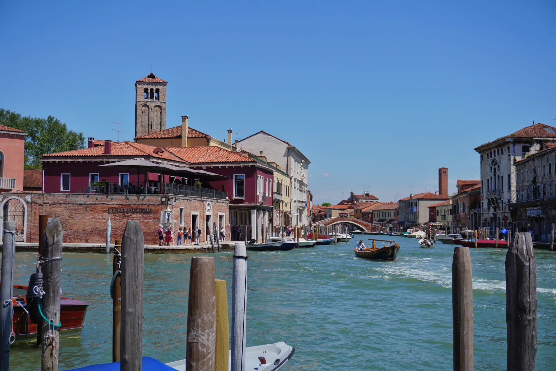 A boatman steers a gondola down a shimmering turquoise canal in Venice, Italy, with ornate historic buildings and terracotta roofs lining the banks under a clear blue sky.