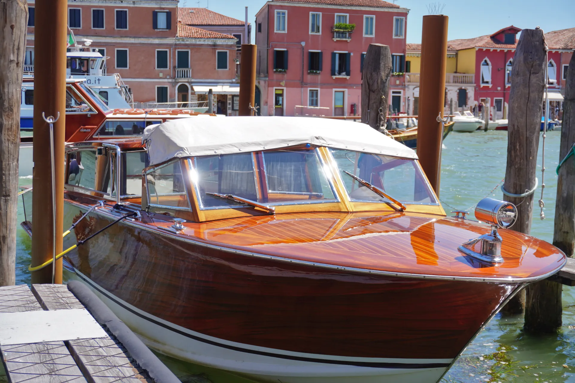 A gleaming wooden boat is tied to wooden posts in a canal on Murano island, Venice, Italy. Colorful buildings line the waterway in the background.