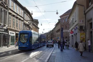 A blue tram moves down a street in Zagreb, Croatia, with shops and historic buildings on either side and pedestrians on the sidewalk.