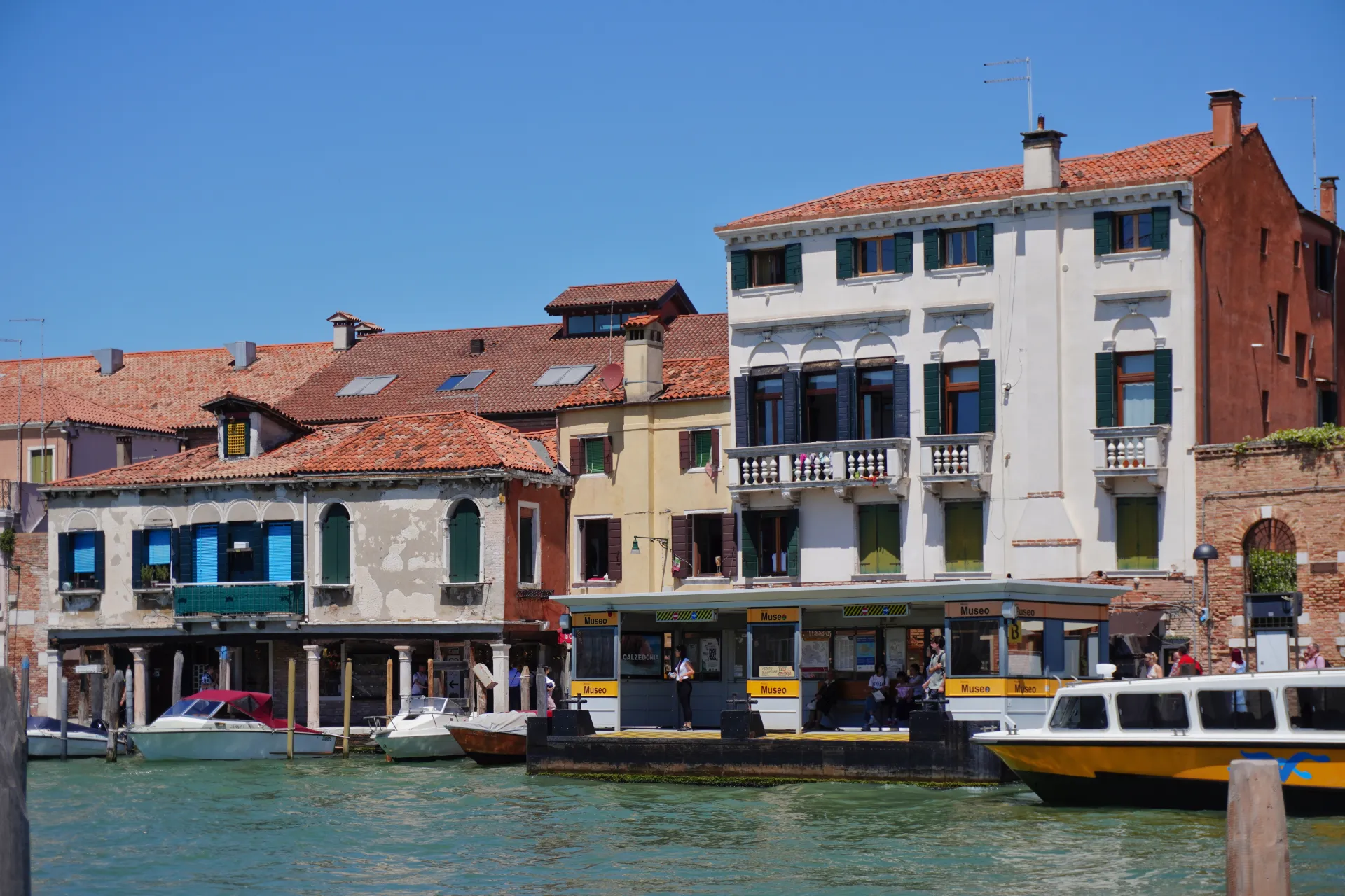 View of a canal in Venice, Italy, with colorful buildings lining the waterway and a water taxi at a dock. A sign reads 'Museo', suggesting this is near the island of Murano.