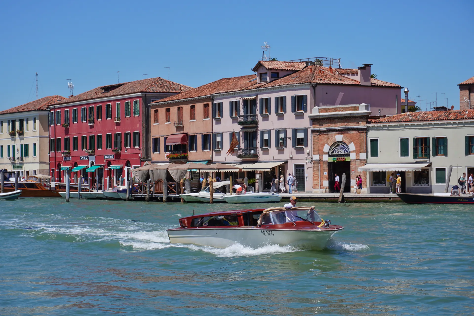 A red and white water taxi speeds down a canal on Murano Island, Venice, Italy, with colorful buildings and docked boats lining the shore under a clear blue sky.