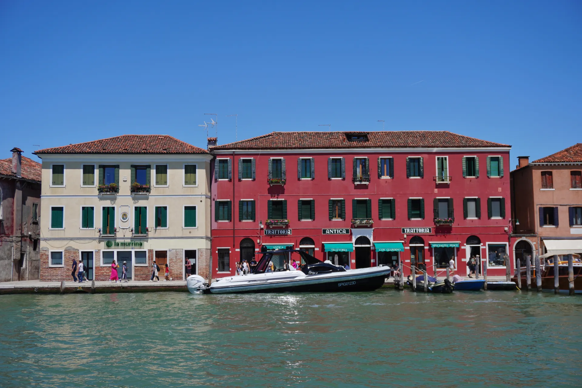 A colorful row of buildings along a Venetian canal, with signs for 'La Murrina', 'Trattoria', and 'Antica'. A motorboat is moored, and people are walking along the waterfront. The architecture evokes the style found on Murano island.