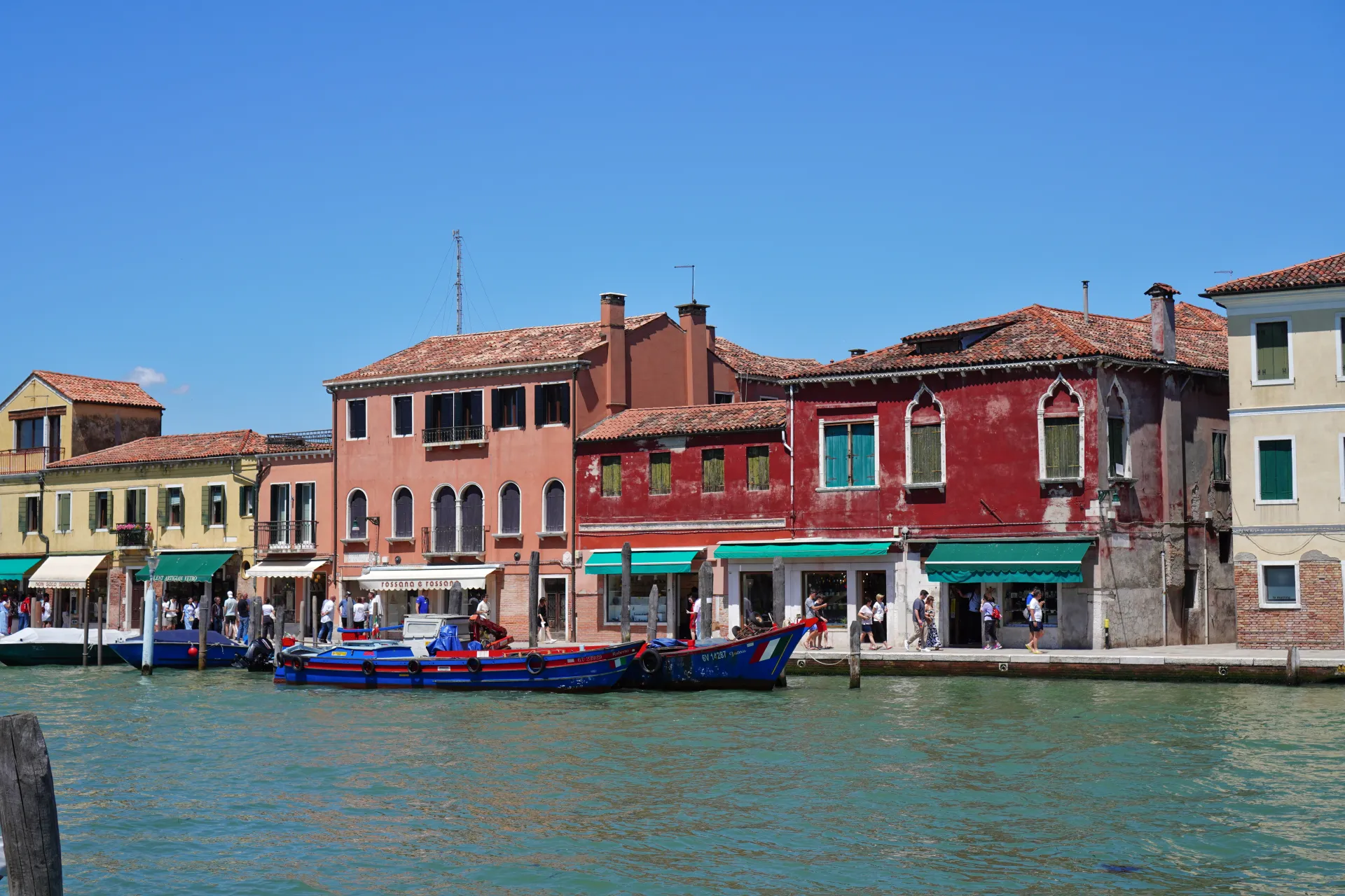 A canal in Venice, Italy, with brightly colored buildings, including red and pink structures, lining the waterfront on Murano island. Boats are docked along the canal, and pedestrians are visible on the walkway in front of shops under a clear blue sky.