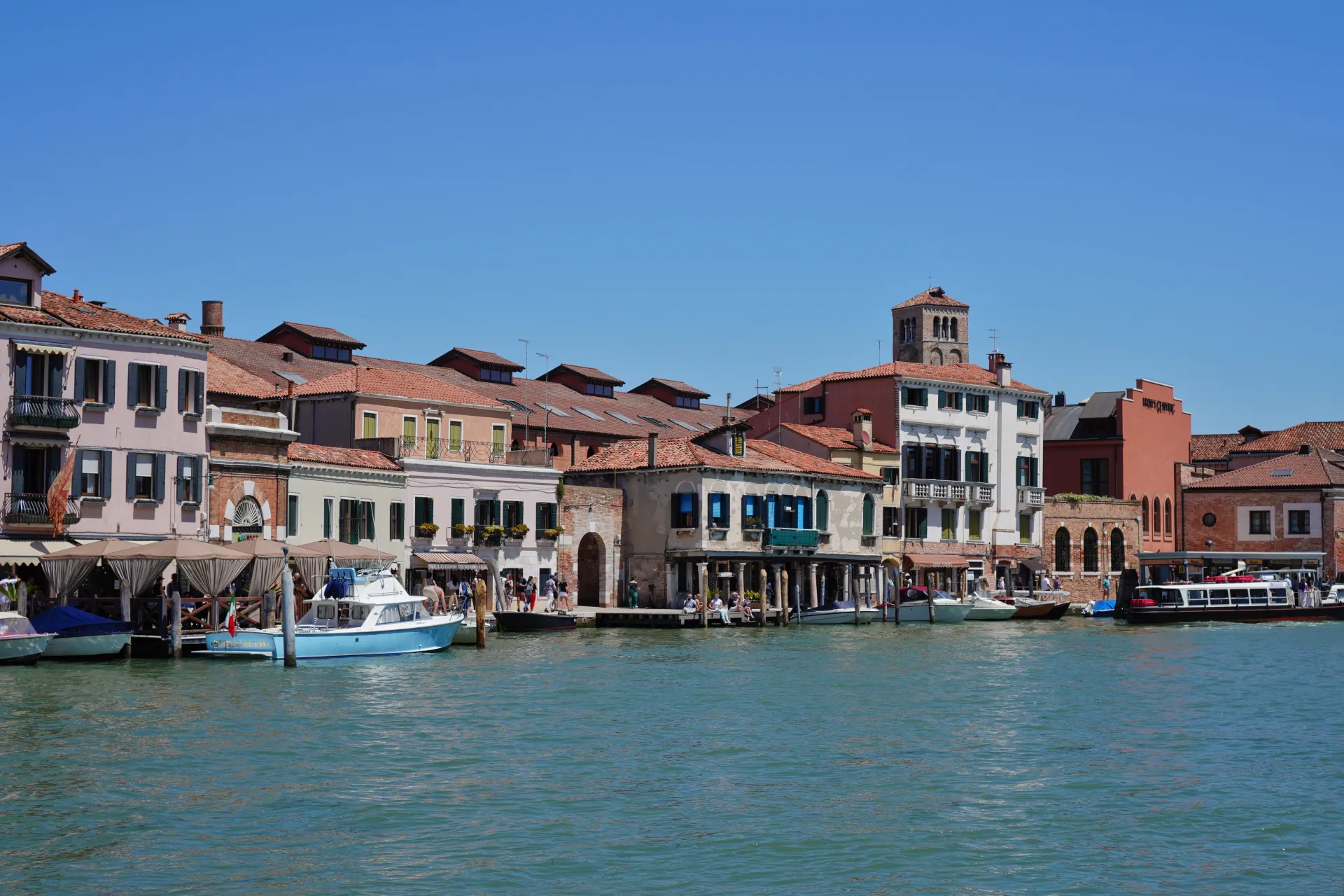 View of the canal on Murano island in Venice, Italy, with traditional buildings, boats docked, and people enjoying the scenery under a clear blue sky.