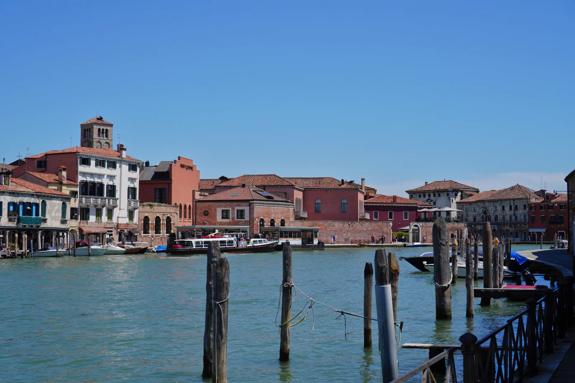 A wide canal in Venice, Italy, with buildings lining the banks. The architecture features red-tiled roofs and colorful facades. Wooden posts and ropes are in the foreground, and several boats, including a passenger ferry, are on the water under a clear blue sky. This scene is characteristic of the island of Murano.