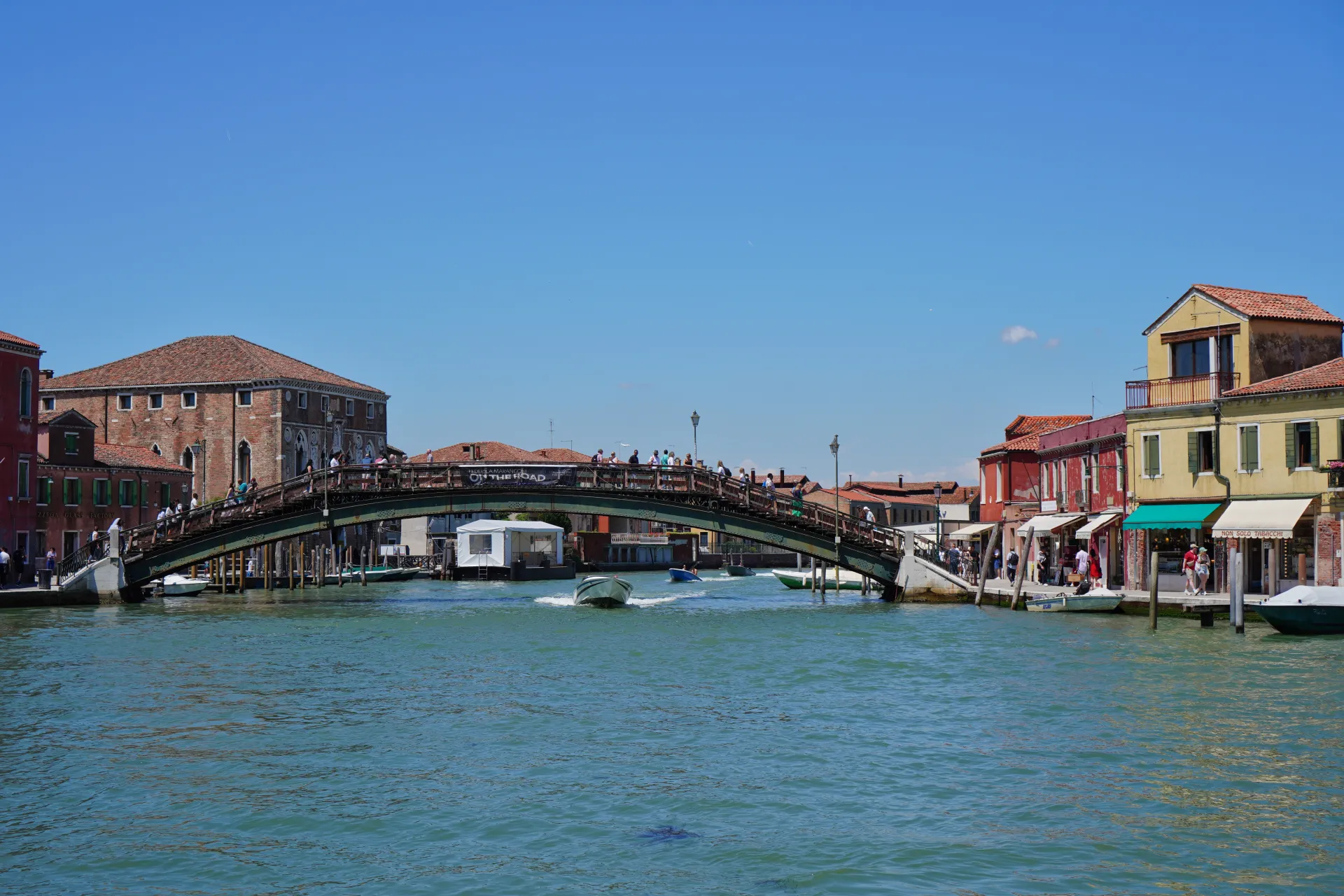 A lively wooden bridge spans a canal on Murano Island, Venice, Italy, with people walking across and boats in the water, under a clear blue sky.