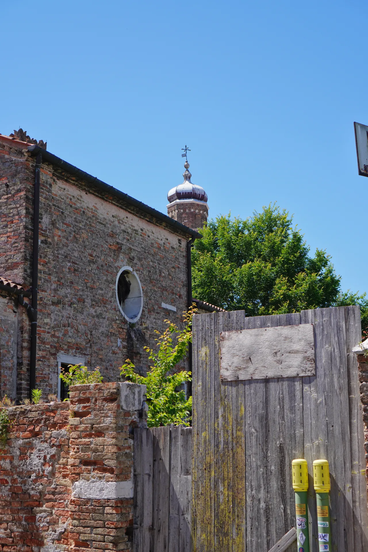A weathered brick building and a rustic wooden fence on Murano island, with a church tower visible in the background under a clear blue sky in Venice, Italy.