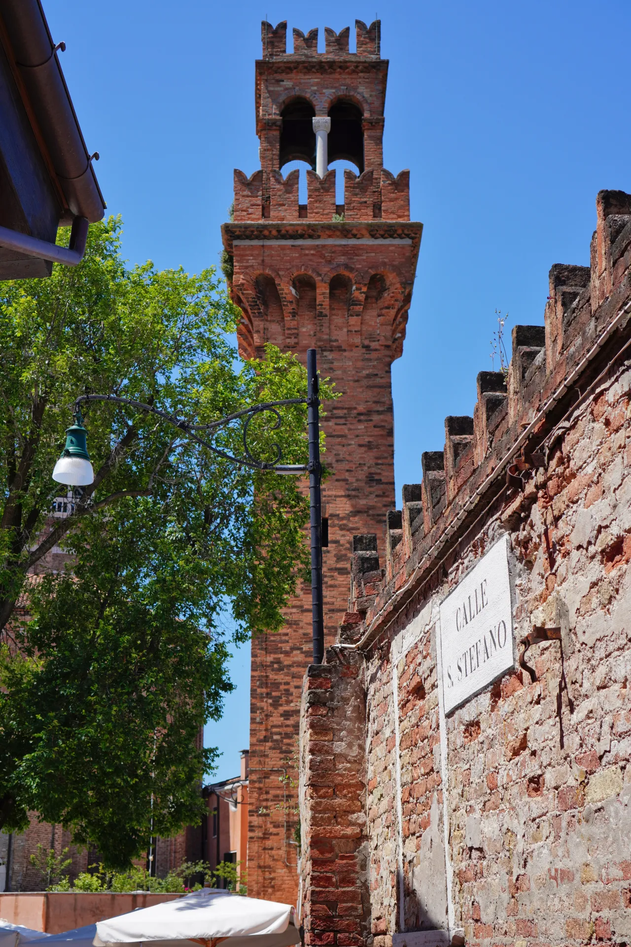 A historic red brick tower with battlements stands tall against a bright blue sky on the island of Murano, Venice. A sign for 'Calle S. Stefano' is on a weathered brick wall in the foreground, with a tree and street lamp adding to the scene.