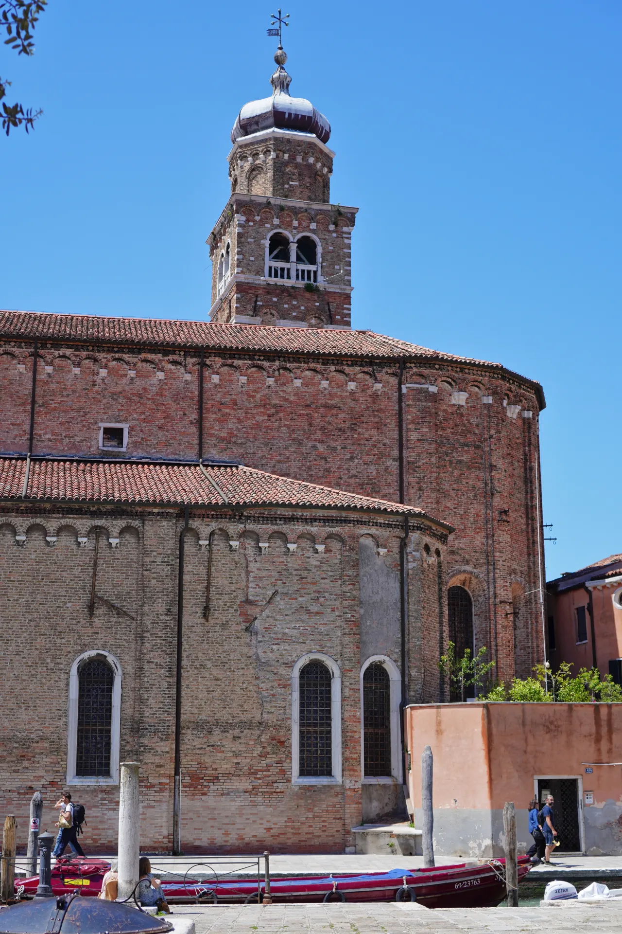 A brick church with a multi-tiered bell tower stands under a clear blue sky on Murano island, Venice. Red tiled roofs and arched windows are visible, with canal boats in the foreground.