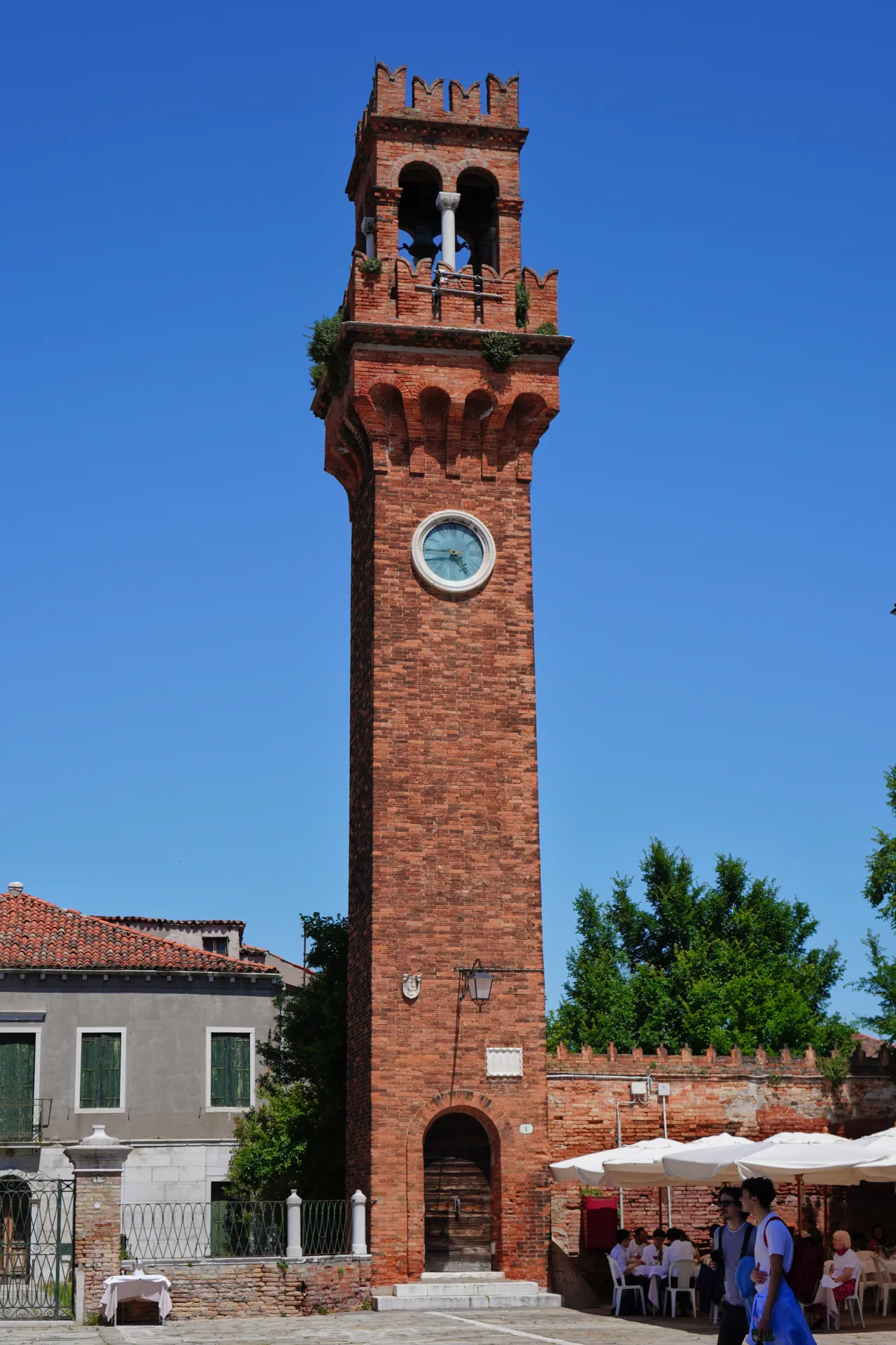 The red brick clock tower on the island of Murano, Venice, Italy, under a bright blue sky. The tower has a clock face and decorative battlements.