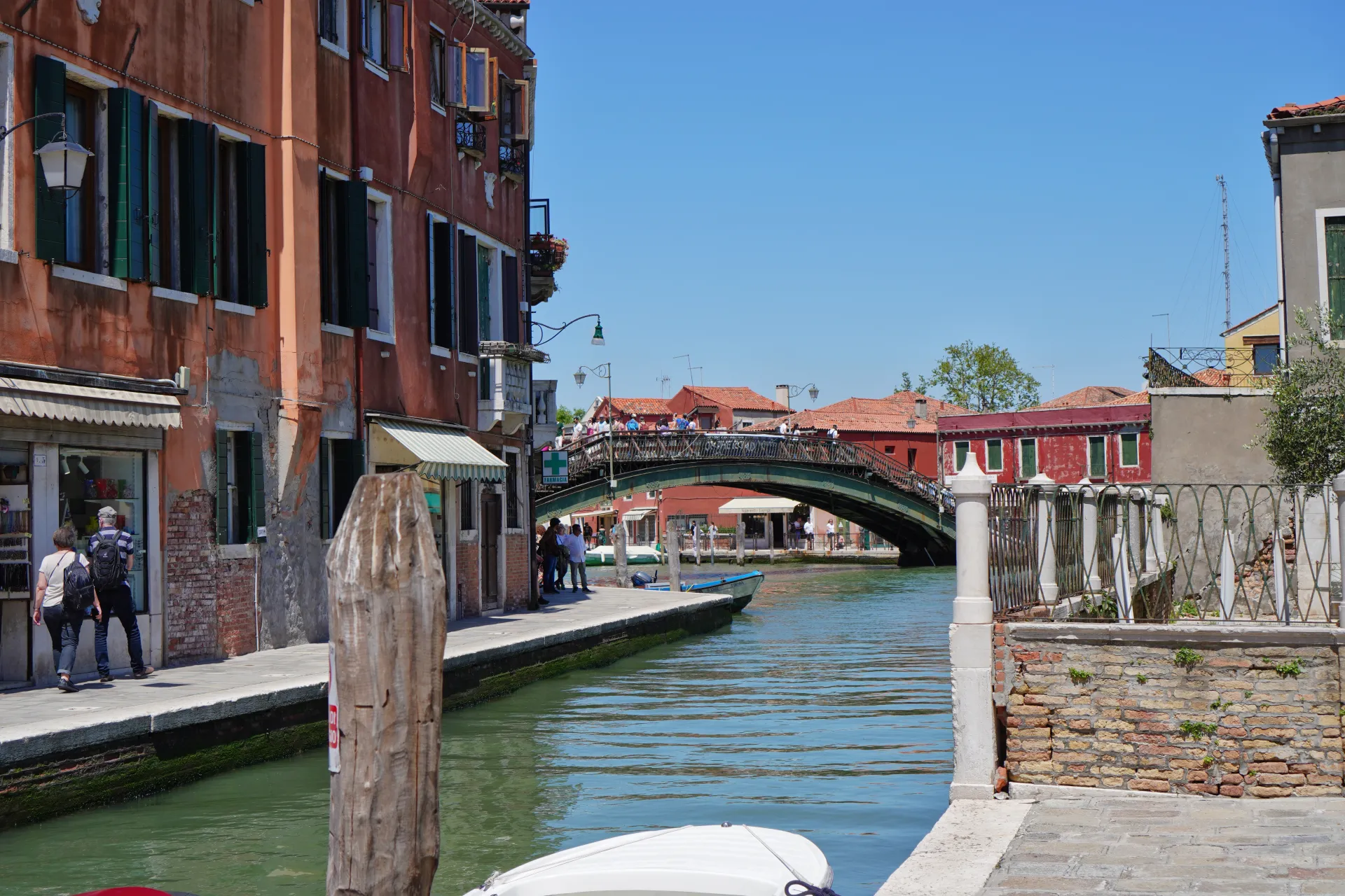 A serene canal in Venice, Italy, with old buildings lining the waterway. A metal pedestrian bridge arches over the canal, crowded with people. Boats are moored along the embankment. The scene carries the distinct charm associated with Venice and hints at the tranquil atmosphere of the island of Murano.
