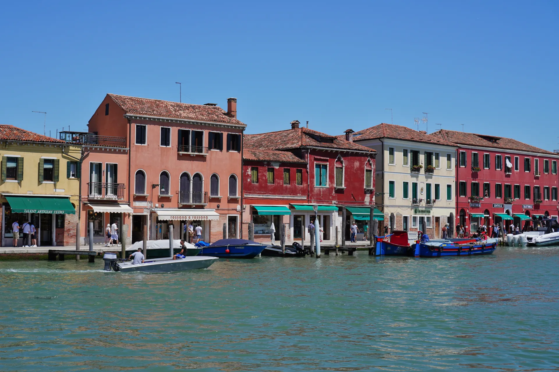 Pogled na šarene zgrade i čamce duž kanala na ostrvu Murano, Venecija, Italija, pod vedrim plavim nebom.