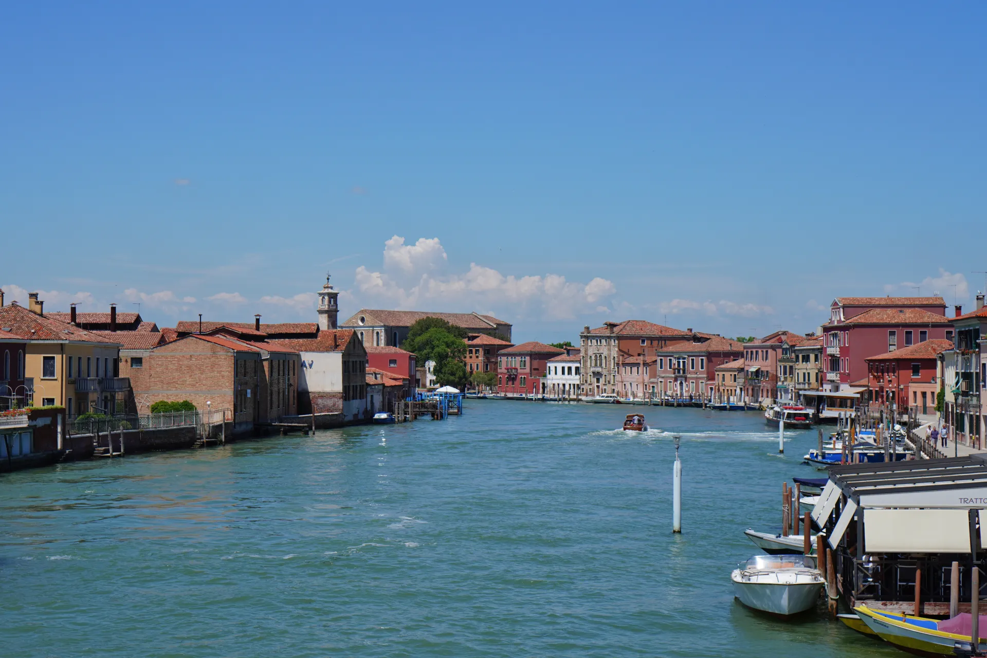 Širok kanal na ostrvu Murano u Veneciji, Italija, sa živopisnim šarenim zgradama i čamcima pod vedrim plavim nebom.