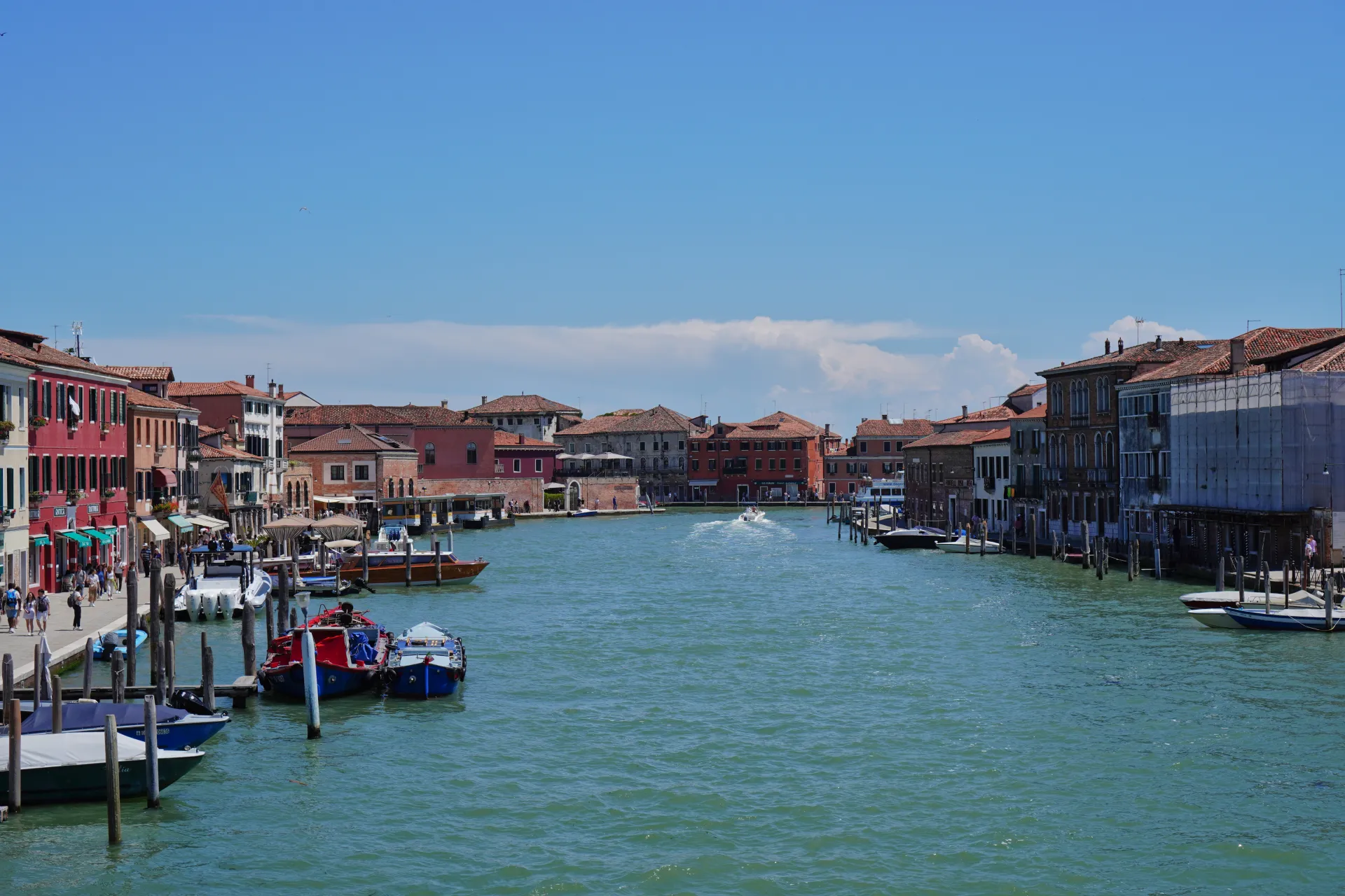 Širok kanal na ostrvu Murano, Venecija, Italija, pod vedrim plavim nebom. Zgrade sa terakotnim krovovima i šarenim fasadama nižu se duž kanala. Mnogi brodovi, uključujući vodene taksije i manje plovile, usidreni su ili se kreću po vodi. Pešaci šetaju duž obalnog šetališta.