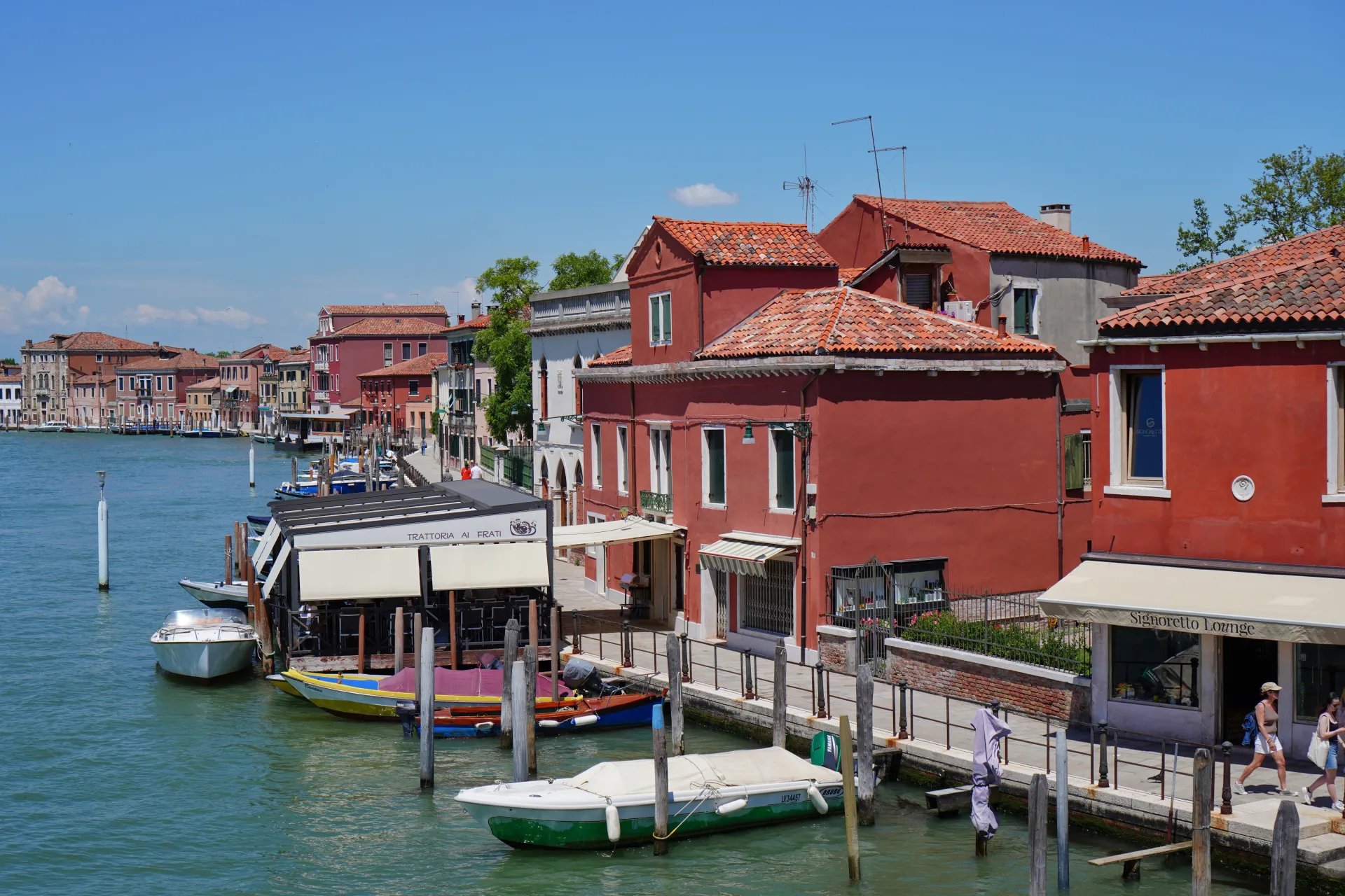 Buildings and boats along the canal on Murano Island, Venice, Italy, under a clear blue sky.