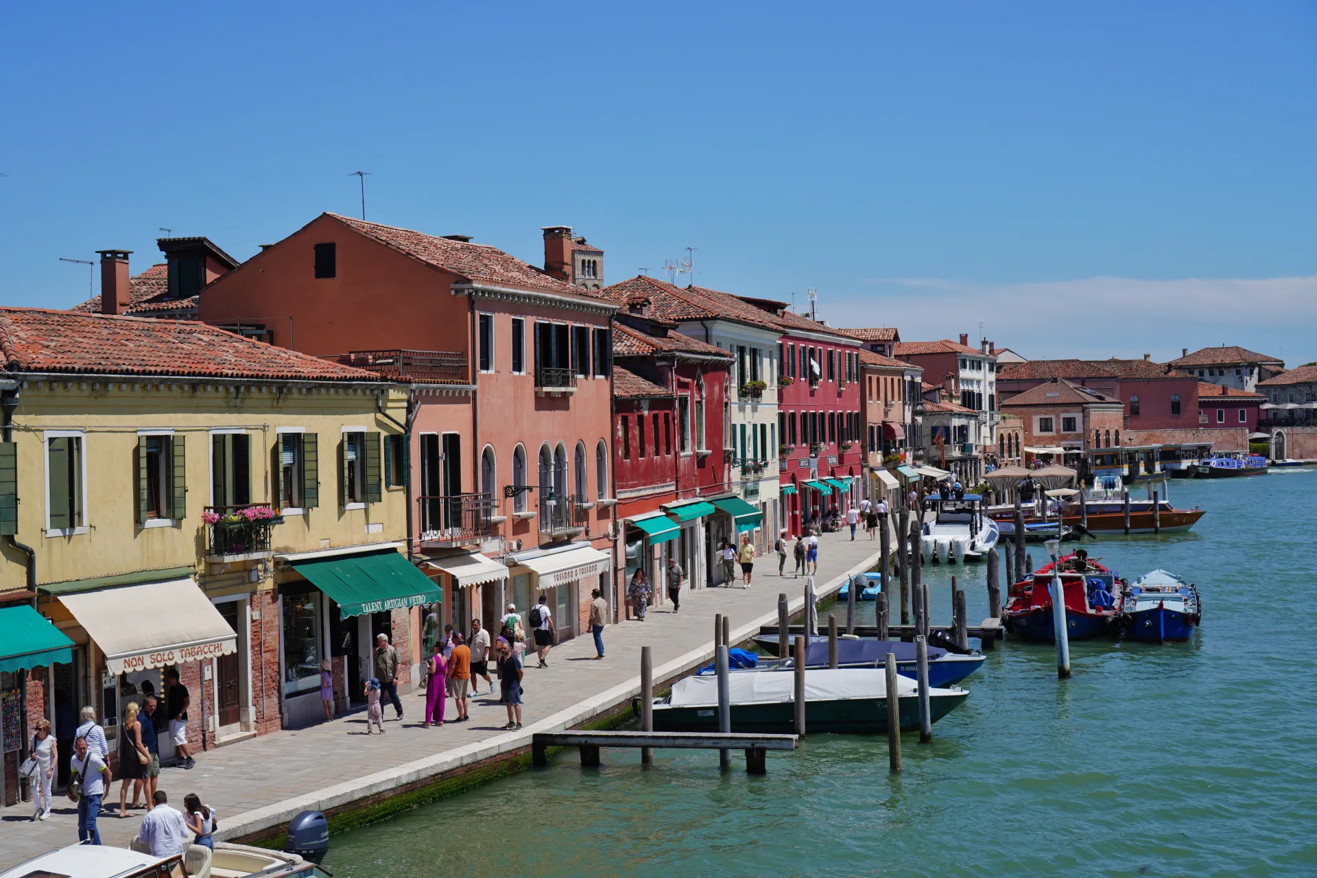 View of colorful buildings lining a canal on Murano island, Venice, Italy, with boats docked in the water and people walking along the waterfront.