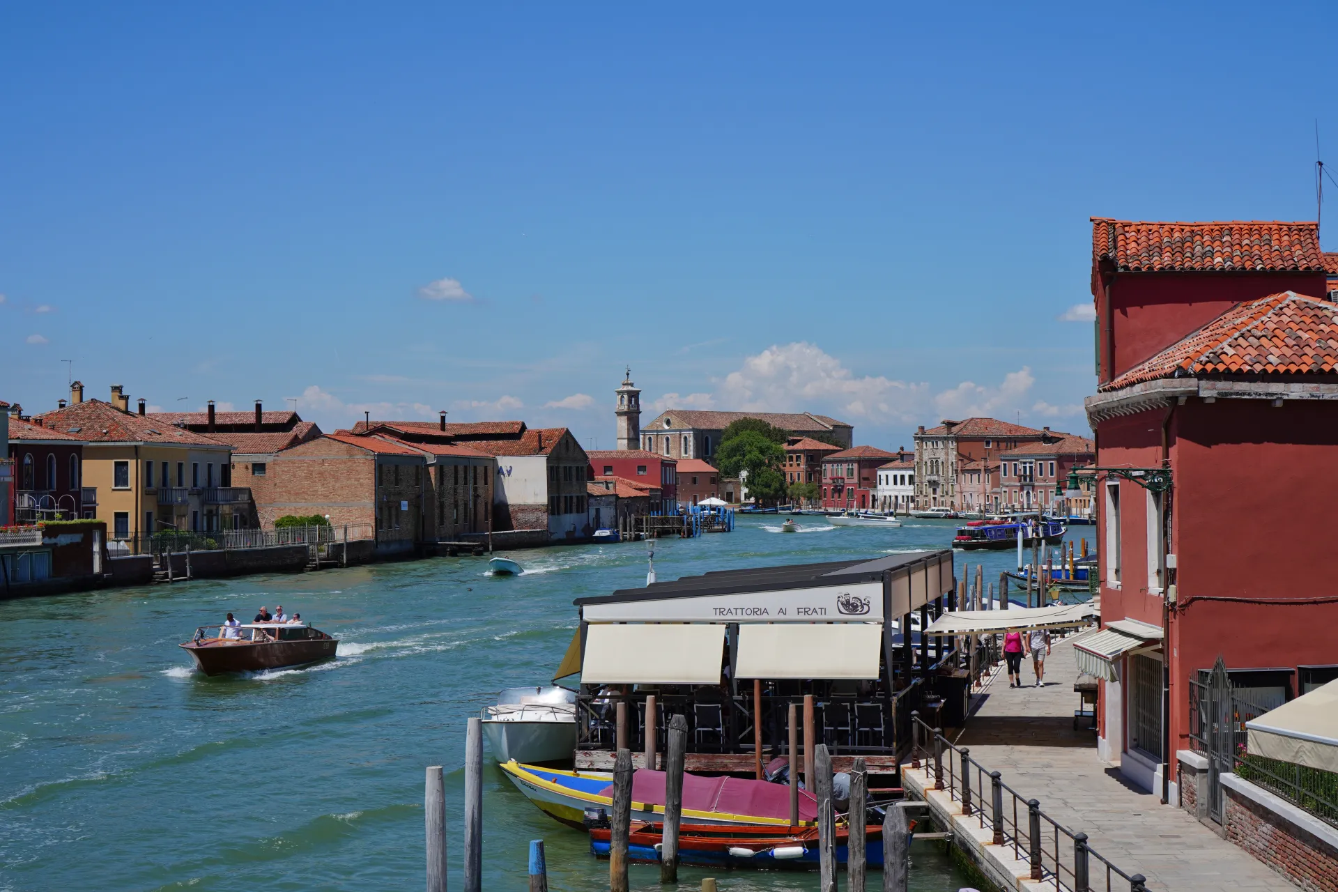 A boat speeds down a canal on Murano Island, Venice, Italy, with colorful buildings lining the banks and a trattoria on the right.
