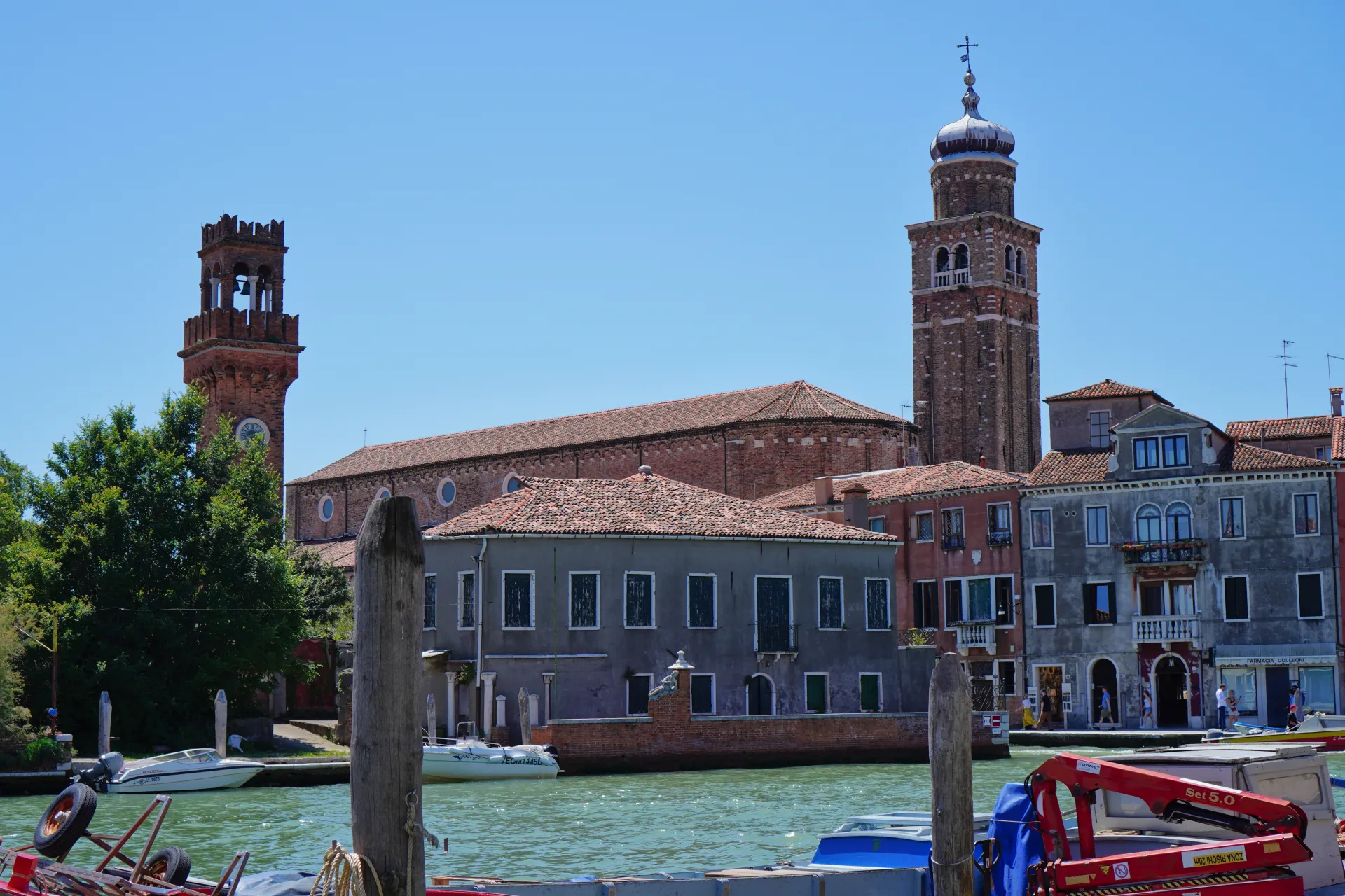 A canal on Murano island, Venice, Italy, with ancient brick bell towers, tiled roofs, and buildings lining the waterway under a clear blue sky.