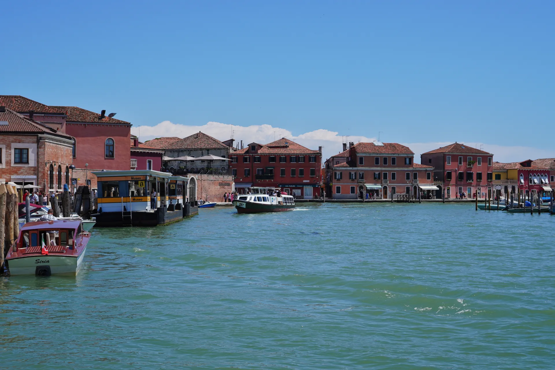 Waterfront buildings and boats on the canal of Murano island, Venice, Italy under a clear blue sky.