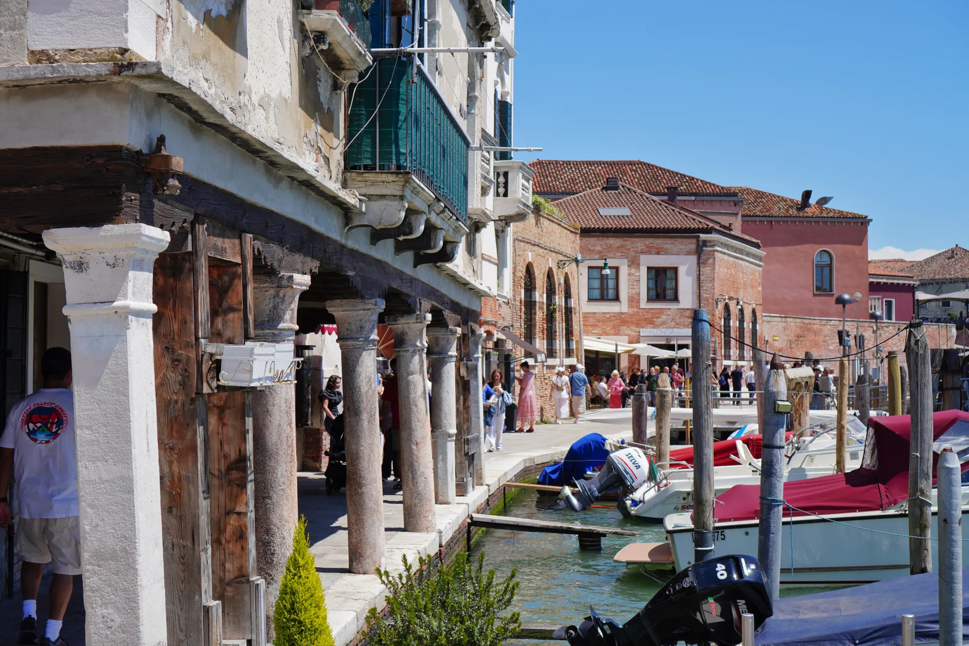 A canal lined with old buildings and boats on Murano Island in Venice, Italy, with people walking along the promenade.
