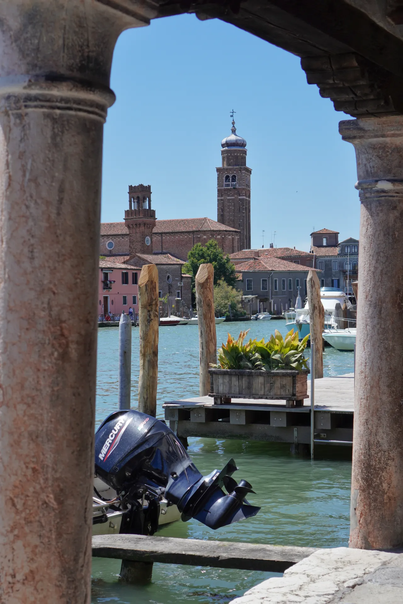 View of a canal on Murano island, Venice, with a motorboat in the foreground and historic buildings and bell towers in the background, framed by ancient stone columns.