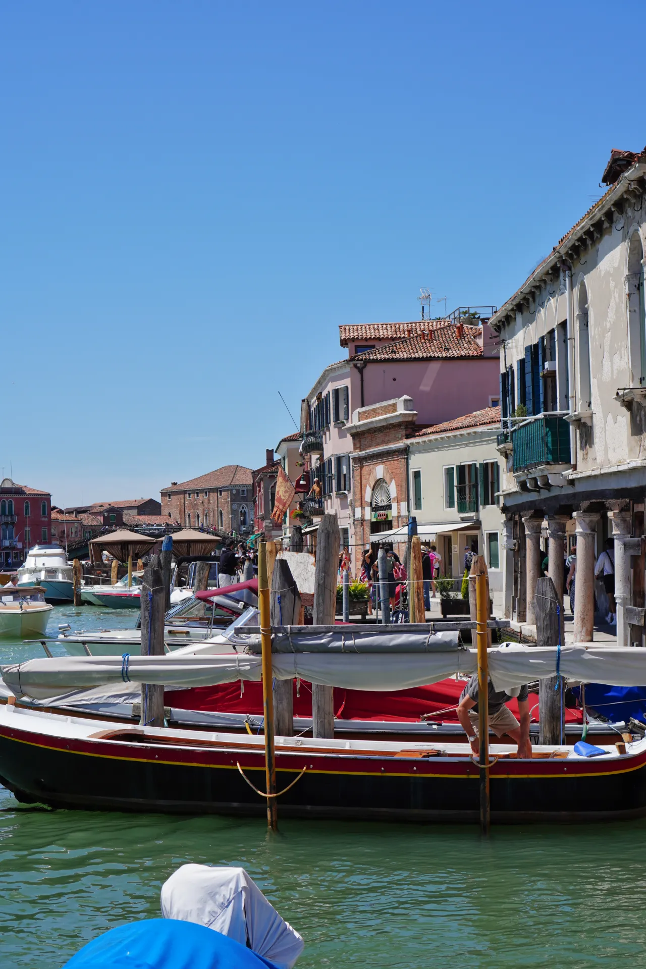 Traditional boats are moored along a canal on the island of Murano in Venice, Italy, with colorful buildings and people on the waterfront under a clear blue sky.