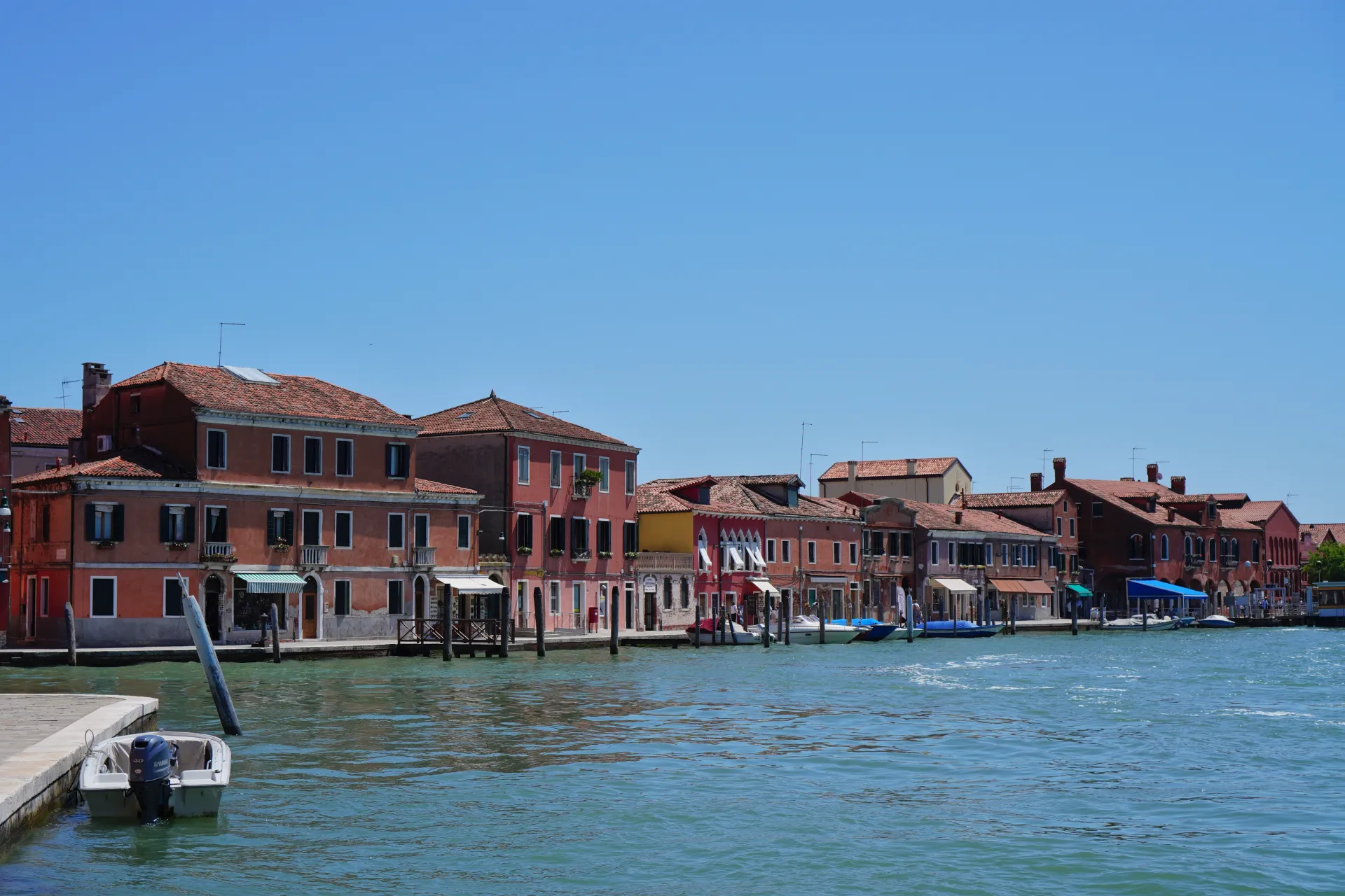 View of colorful historic buildings on Murano Island, Venice, Italy, with canals and boats in the foreground under a bright blue sky.