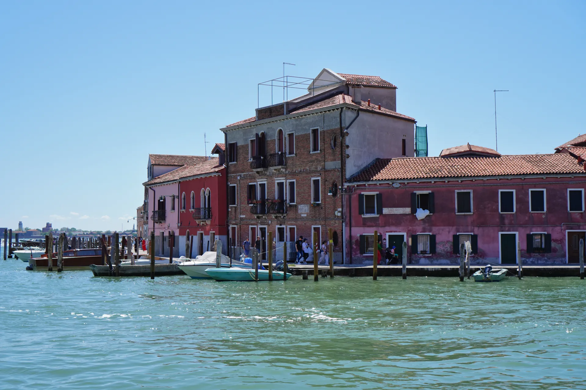 Waterfront buildings with colorful facades on Murano Island, Venice, Italy, with boats moored in the canal and people walking along the path.